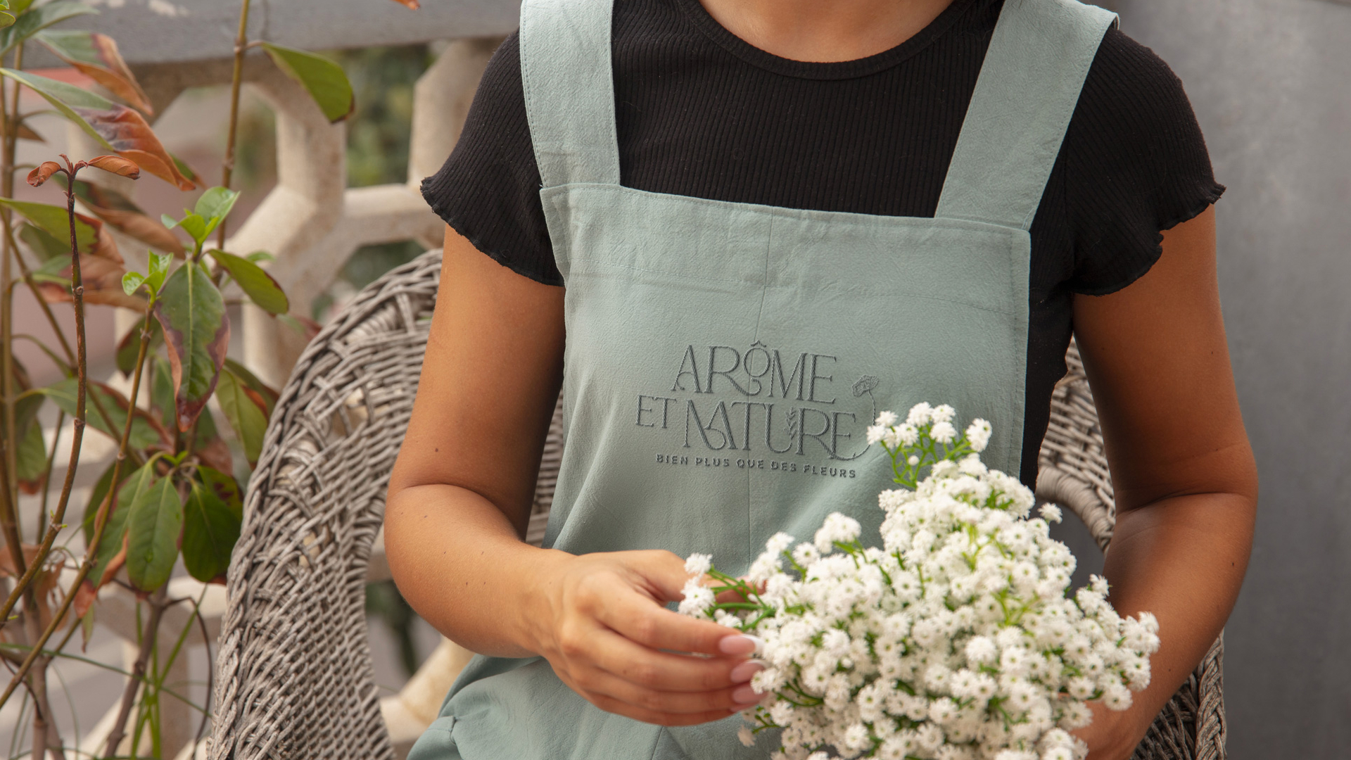 Fleuriste portant un tablier vert “Arôme et Nature” et tenant un bouquet de petites fleurs blanches.