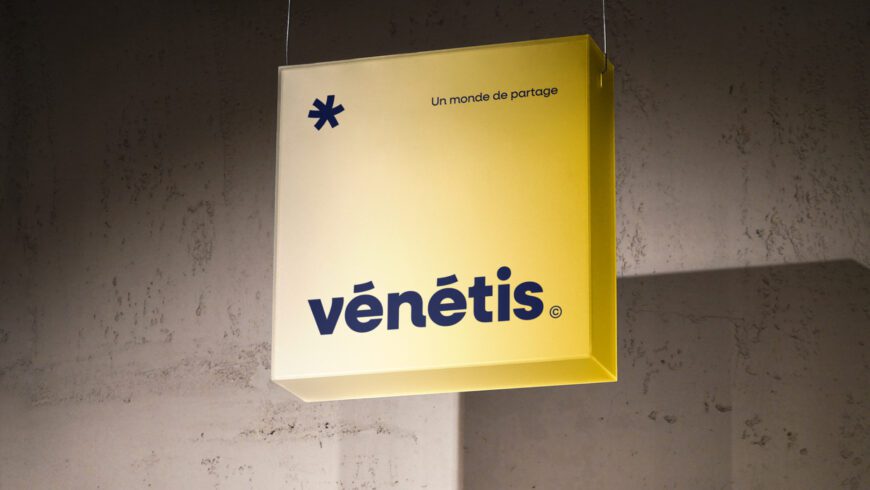 Enseigne lumineuse jaune présentant le nouveau logo Vénétis et le slogan « Un monde de partage », conçue par Pois Chiche Design à Lorient dans le cadre de la refonte d’identité visuelle.