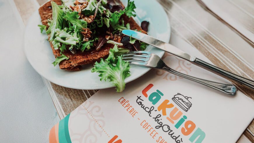 Assiette de galette bretonne avec salade, posée à côté du menu coloré du restaurant Lã Kuign à Lorient, identité visuelle signée Pois Chiche Design.