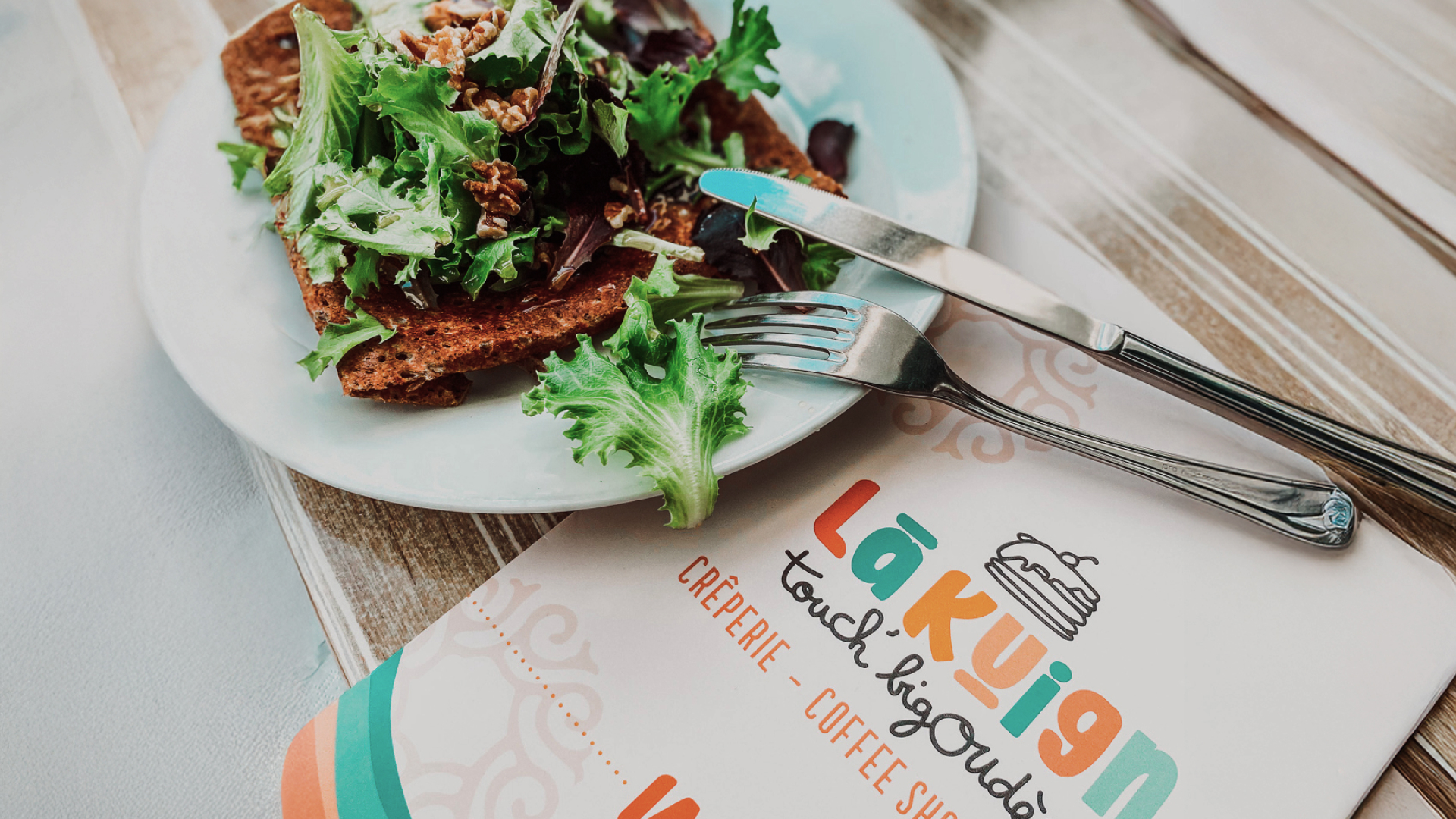 Assiette de galette bretonne avec salade, posée à côté du menu coloré du restaurant Lã Kuign à Lorient, identité visuelle signée Pois Chiche Design.